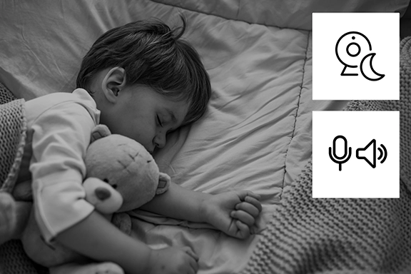 Night shot of a toddler sleeping in bed. The icons for night vision and microphone are shown in the top right-hand corner, indicating that the camera can be used as a baby monitor