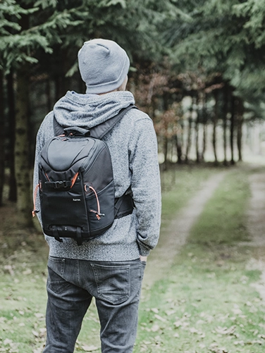 A man stands in front of a forest and looks into it, carrying the Hama camera backpack "Profitour" on his back